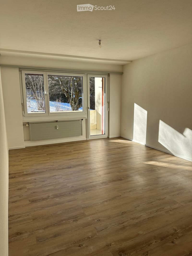 Empty room with wooden flooring, large windows, and a radiator. Sunlight streams in from the windows, casting shadows on the floor.