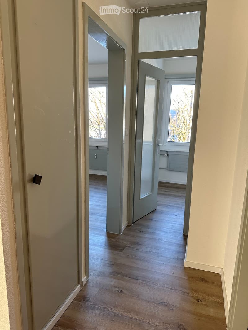 Empty hallway with wooden flooring, light-colored walls, and two open doors leading to adjacent rooms with windows.