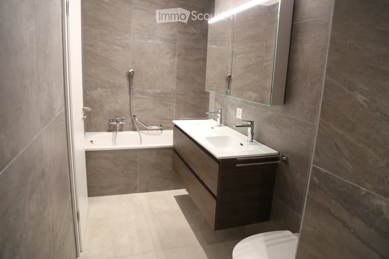 Modern bathroom with a bathtub, double sink, and a large mirror above the vanity. The walls and floor are tiled in a neutral, stone-like pattern.