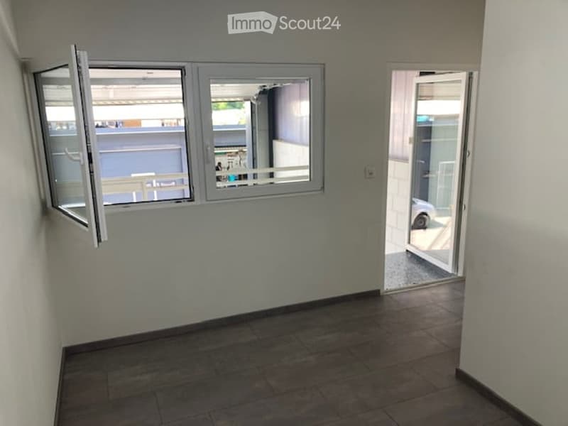Empty room with tiled floor, white walls, and large windows with open shutters. A door leads to an outdoor area.