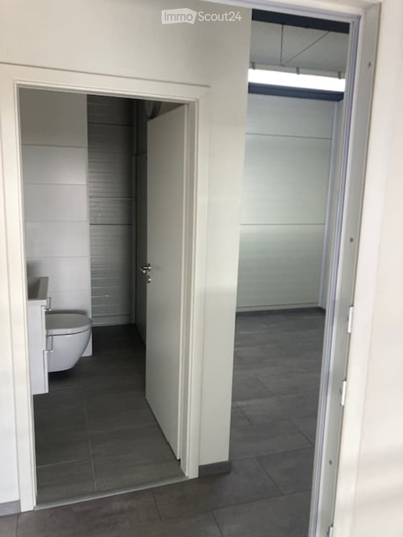 A narrow hallway with white walls and dark tiled flooring, leading to a small bathroom with a toilet and a shower stall.