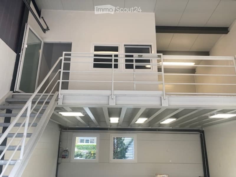 Modern industrial-style building interior with a raised ground floor, metal staircase leading to an upper level, large windows, and exposed ceiling beams.
