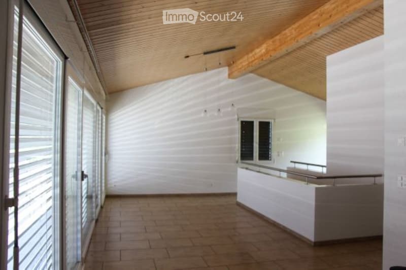 Empty room with white walls, wooden ceiling beams, tiled floor, and large glass windows with blinds. A white railing is visible on the right side.