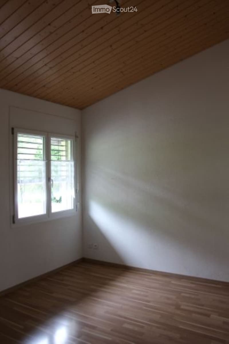 Empty attic room with wooden slanted ceiling, wooden floor, and a single window letting in natural light.