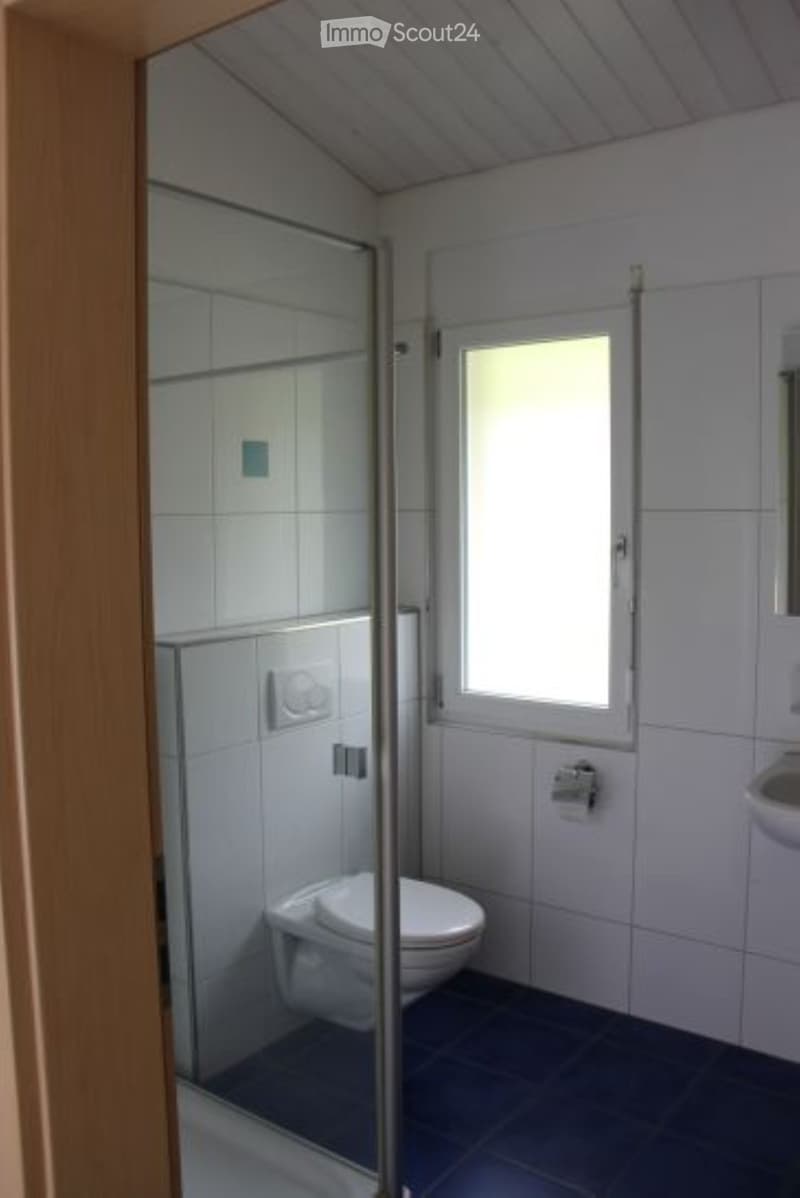 Modern bathroom with white tiles, a toilet, a sink, and a glass-enclosed shower area. The room features a small window for natural light.
