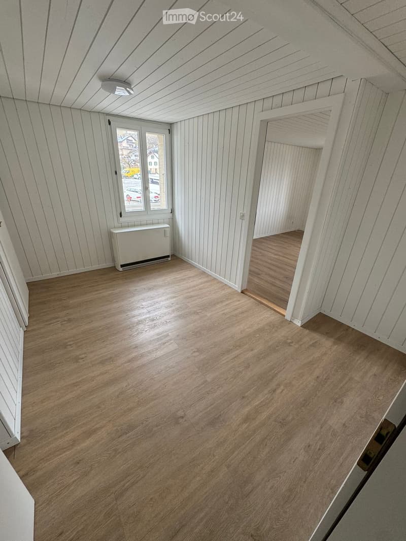 The room has a wooden floor, white walls, a white ceiling, a window with white frames, and a white radiator. There is a doorway that leads to another room with a similar interior.