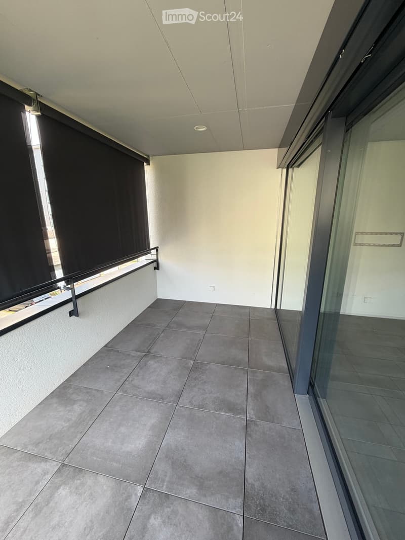 Modern balcony with grey tiled flooring, white walls, and a black metal railing. Features large glass sliding doors and a retractable black blind on one side.