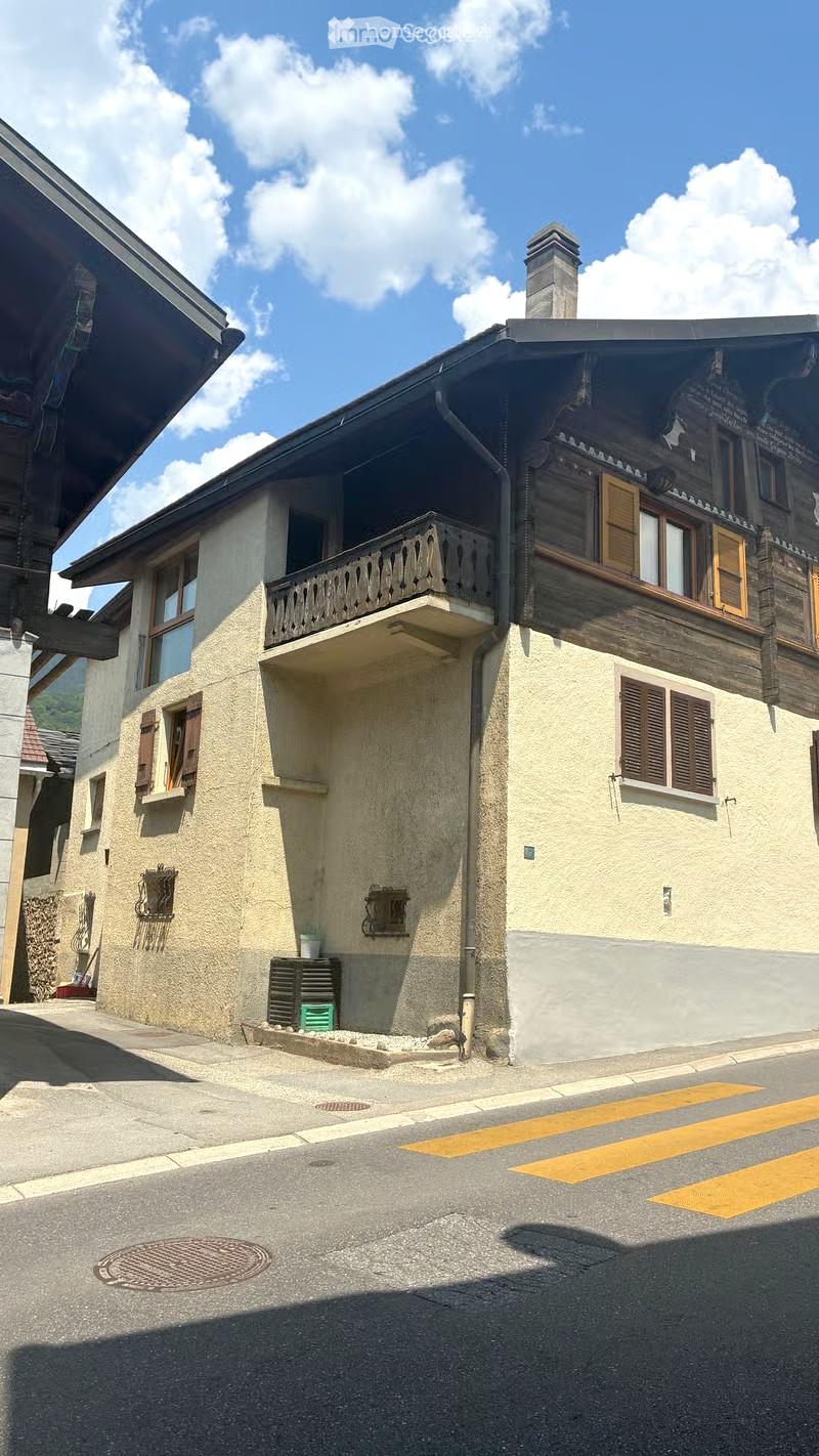 Two-story building with a balcony, wooden shutters, and a chimney. The lower level has a textured beige facade, while the upper level features wooden beams and a decorative balcony railing.
