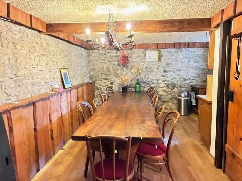 A rustic dining area with a long wooden table surrounded by chairs, a chandelier hanging from the ceiling, and a stone wall backdrop. There is a picture frame on a wooden counter and a vase with flowers on the table.
