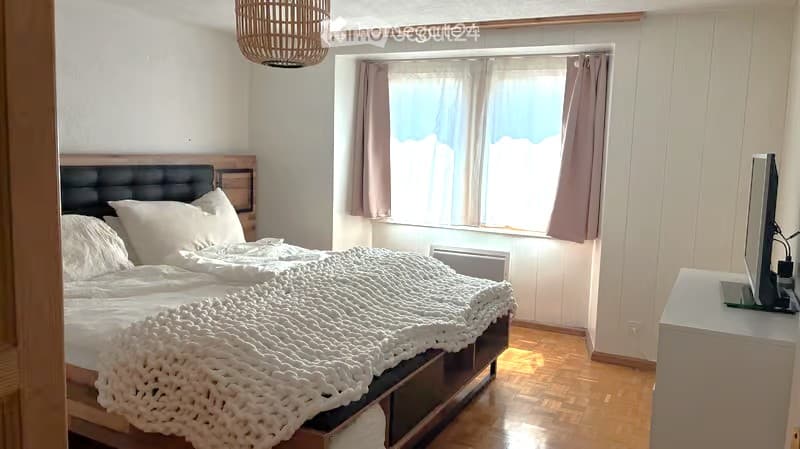 Cozy bedroom with a large bed, white bedding, a nightstand, a screen/monitor, and a window with curtains.