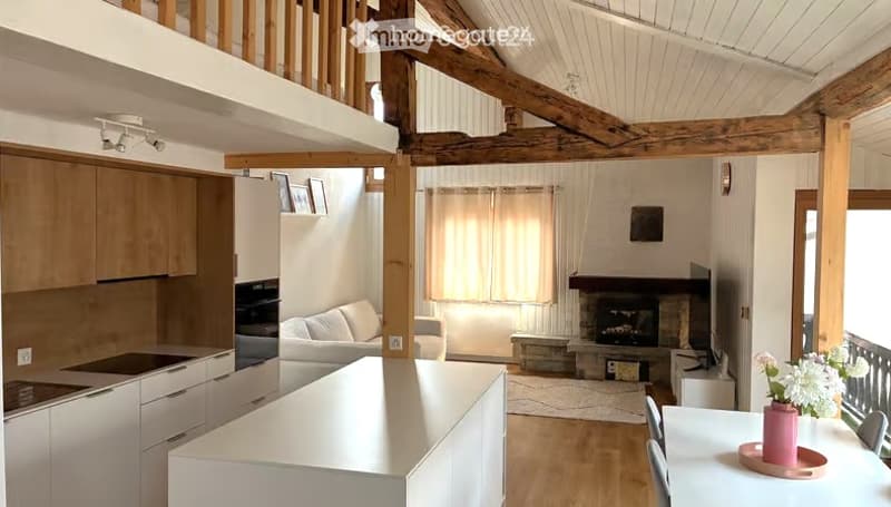 Open-plan living and dining area with a modern kitchen, white cabinets, a central island, a fireplace, and a sofa. Wooden beams on the ceiling and large windows provide natural light.