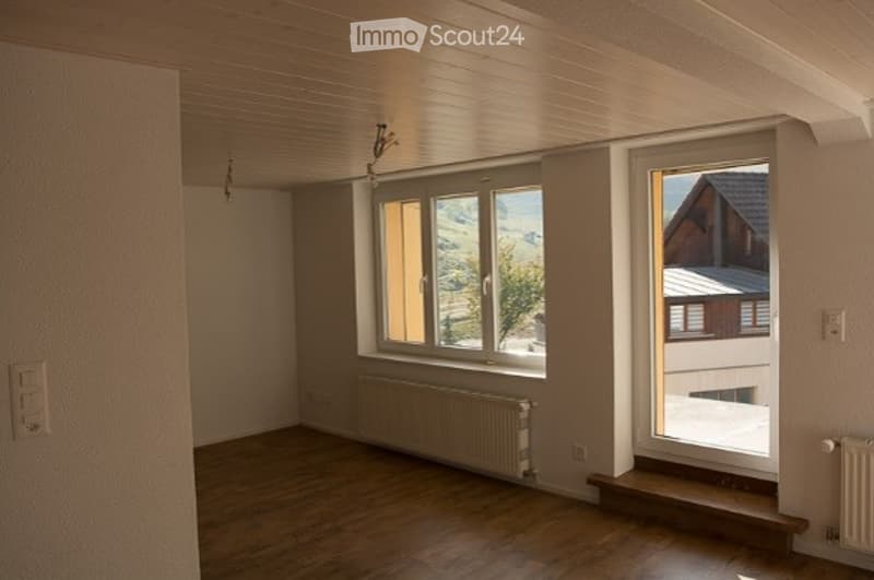 Empty room with wooden flooring, white walls, large windows, radiator, and a small step leading to an outdoor area.