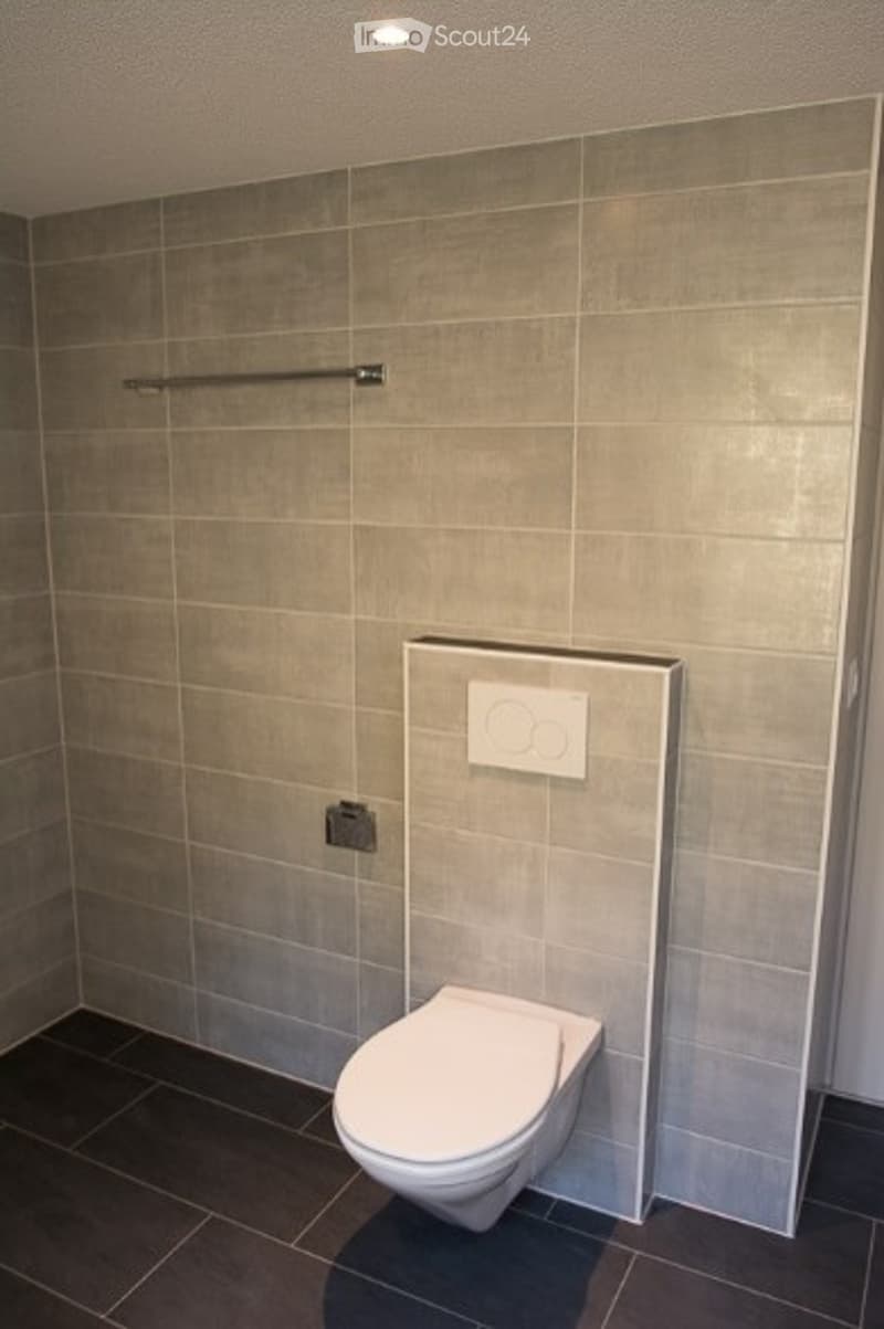 Modern bathroom with grey tiled walls and dark tiled floor, featuring a white toilet and a built-in control panel.