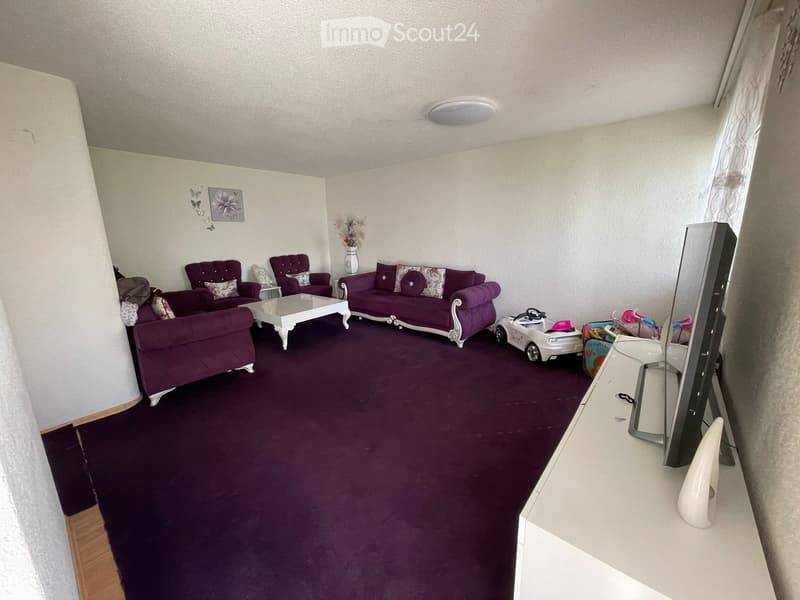 Living room with purple carpeting, multiple purple sofas, a white coffee table, a TV screen, and decorative items including a vase and a toy car.