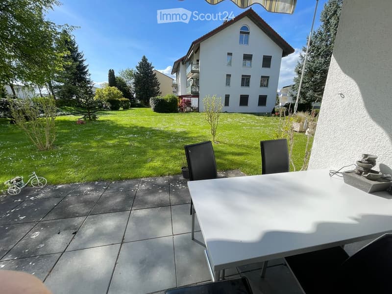 Outdoor patio with a white table and two black chairs, overlooking a well-maintained lawn and a multi-story residential building.