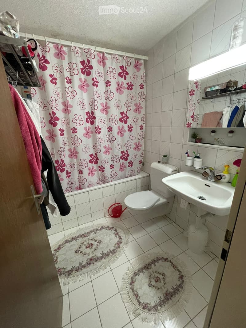 A small bathroom with white tiled walls and floor, featuring a toilet, sink, bathtub with a floral shower curtain, and two round rugs on the floor. A mirror with shelves is mounted above the sink, and a towel hangs on the side.
