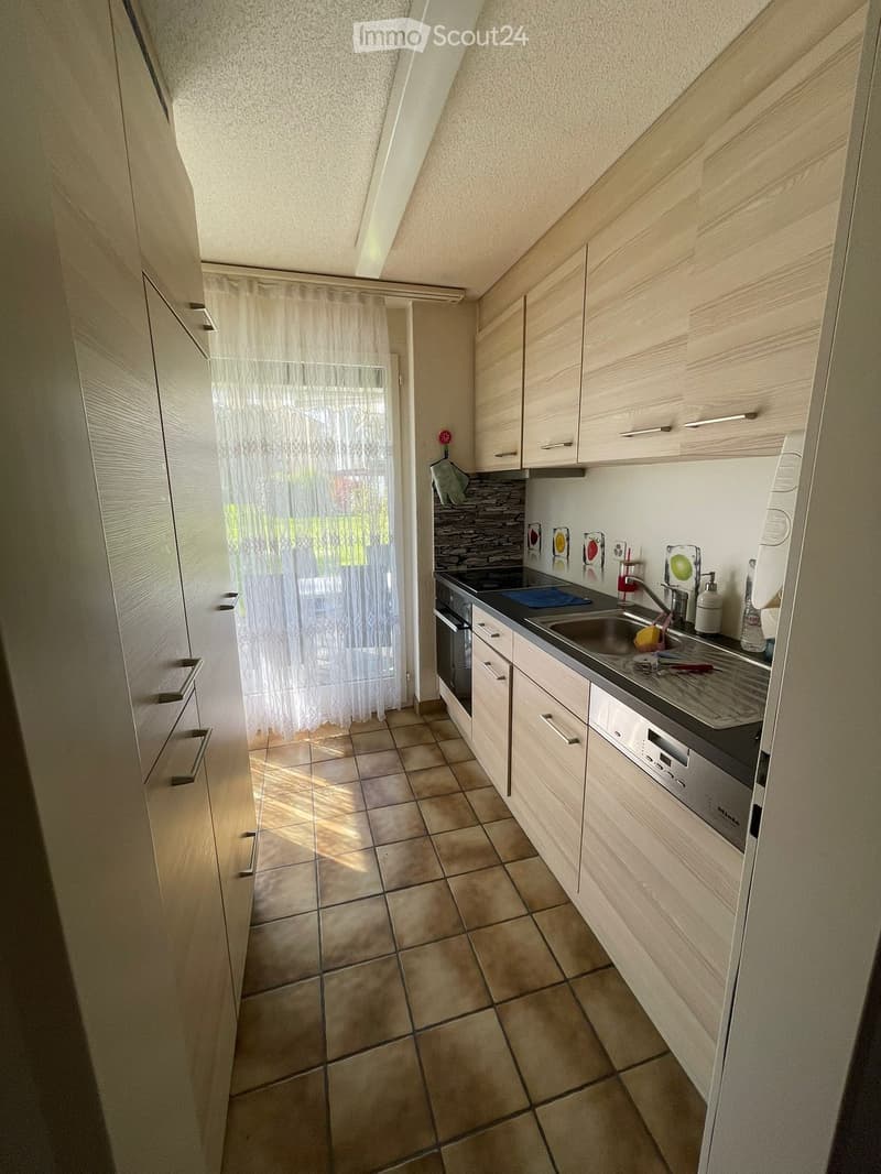 Modern kitchen with light wooden cabinets, tiled floor, and stainless steel appliances including a sink and oven. A curtain covers a window, allowing natural light to enter.