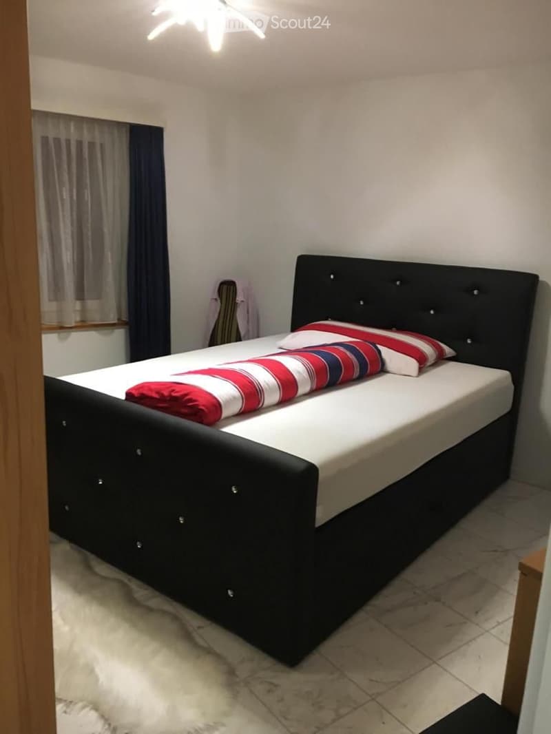 A modern bedroom with a black upholstered bed, white bedding, and red and white striped pillows. The room features a large window with sheer curtains and a chandelier on the ceiling.