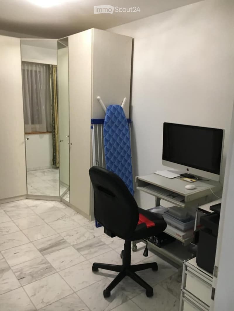 A small, furnished room with a white tiled floor, a black office chair, a white desk with a computer setup, and an ironing board leaning against the wall.
