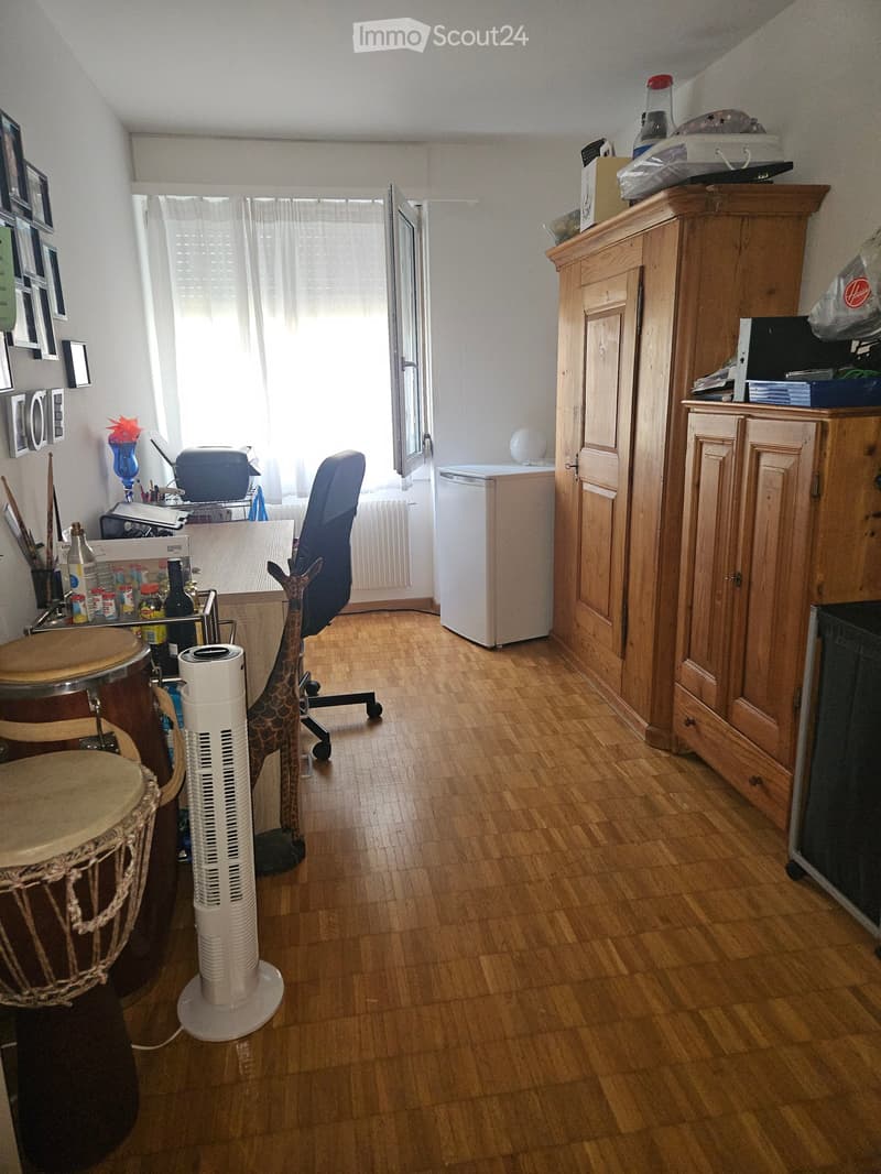 A furnished room with wooden flooring, a large drum, a standing fan, a chair, a refrigerator, and a wooden cabinet.