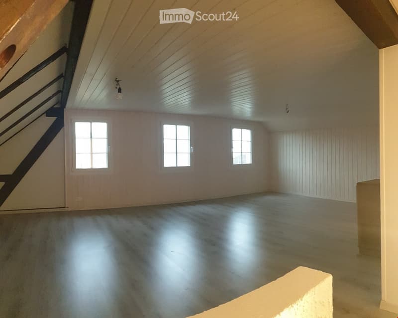 Empty attic space with wooden ceiling, exposed beams, and three windows allowing natural light. The floor is concrete, and there is a staircase leading upwards.