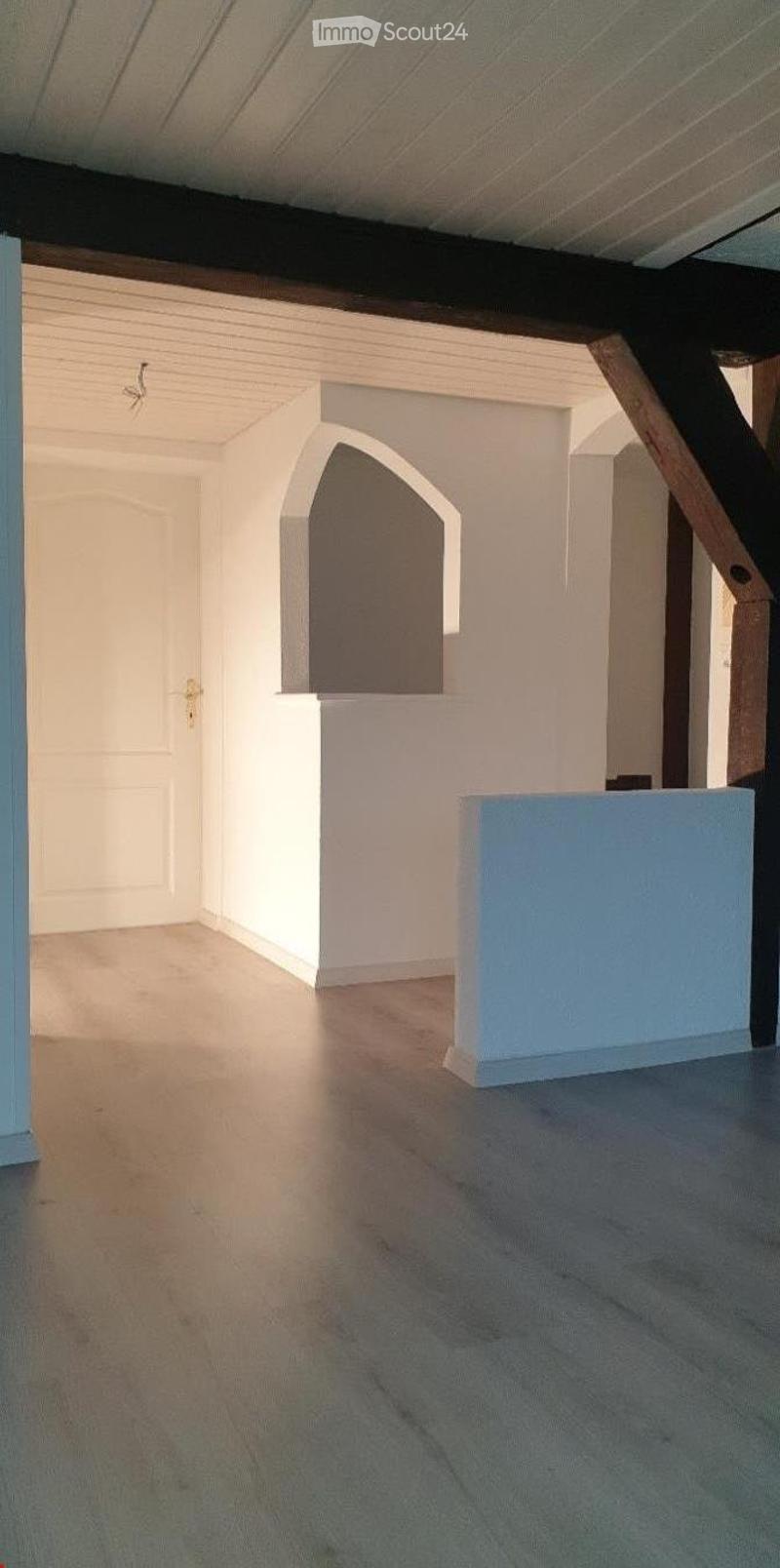 Empty room with wooden flooring, white walls, and a white door. The ceiling has exposed wooden beams and a small arched window.
