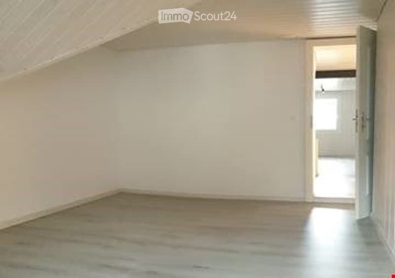 Empty room with light wooden flooring and white walls, featuring a slanted ceiling typical of an attic space. A doorway leads to another room.