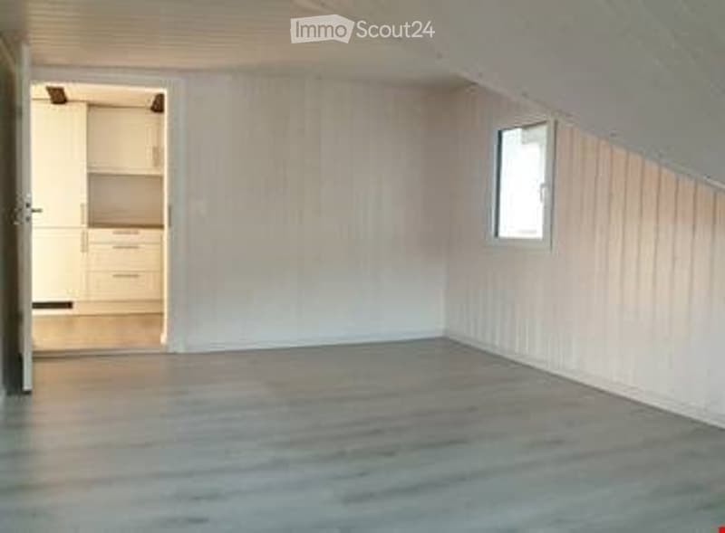 Empty attic room with wooden flooring, white paneled walls, and a small window. No furniture or equipment present.