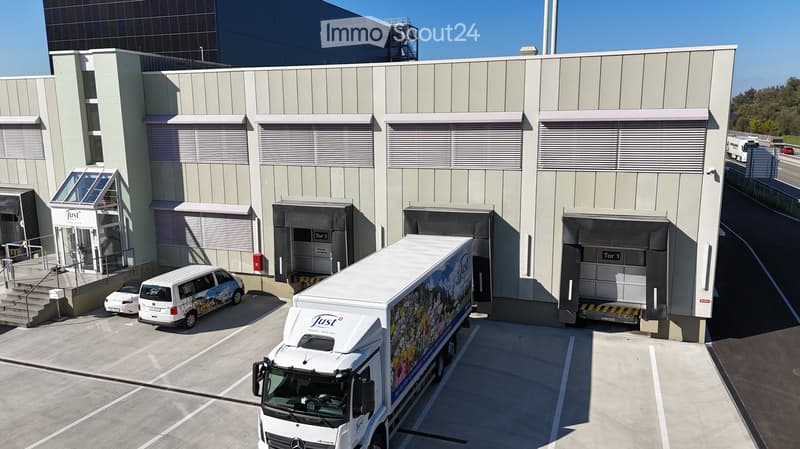 Modern industrial building with multiple loading docks, a large truck parked in front, and a small van and car nearby.