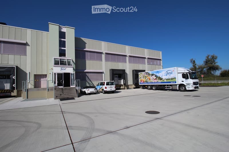 Modern industrial building with a grey facade, multiple loading docks, and a large parking area with several vehicles including a truck and vans.