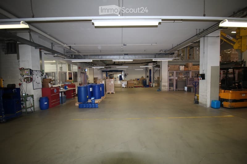 A spacious industrial warehouse with concrete floors, fluorescent lighting, and various storage items including barrels, carts, shelves, and a workbench.