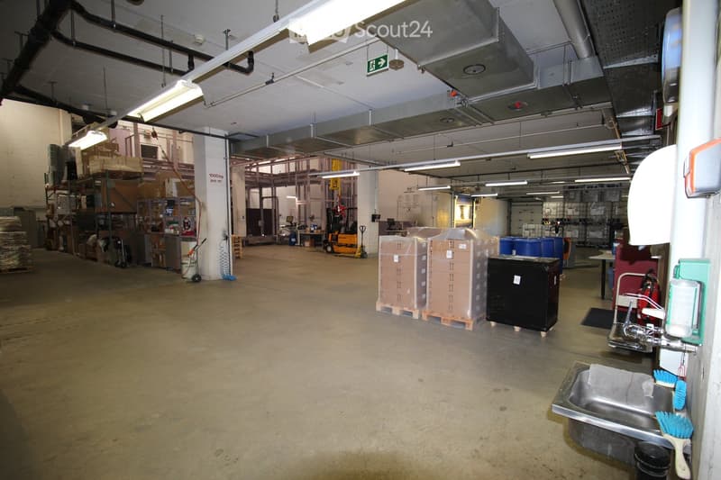 The image shows a large, industrial-style room with a concrete floor and exposed metal beams on the ceiling. The space is well-lit with fluorescent lighting and features various storage items, including stacked cardboard boxes, metal shelving units, and a