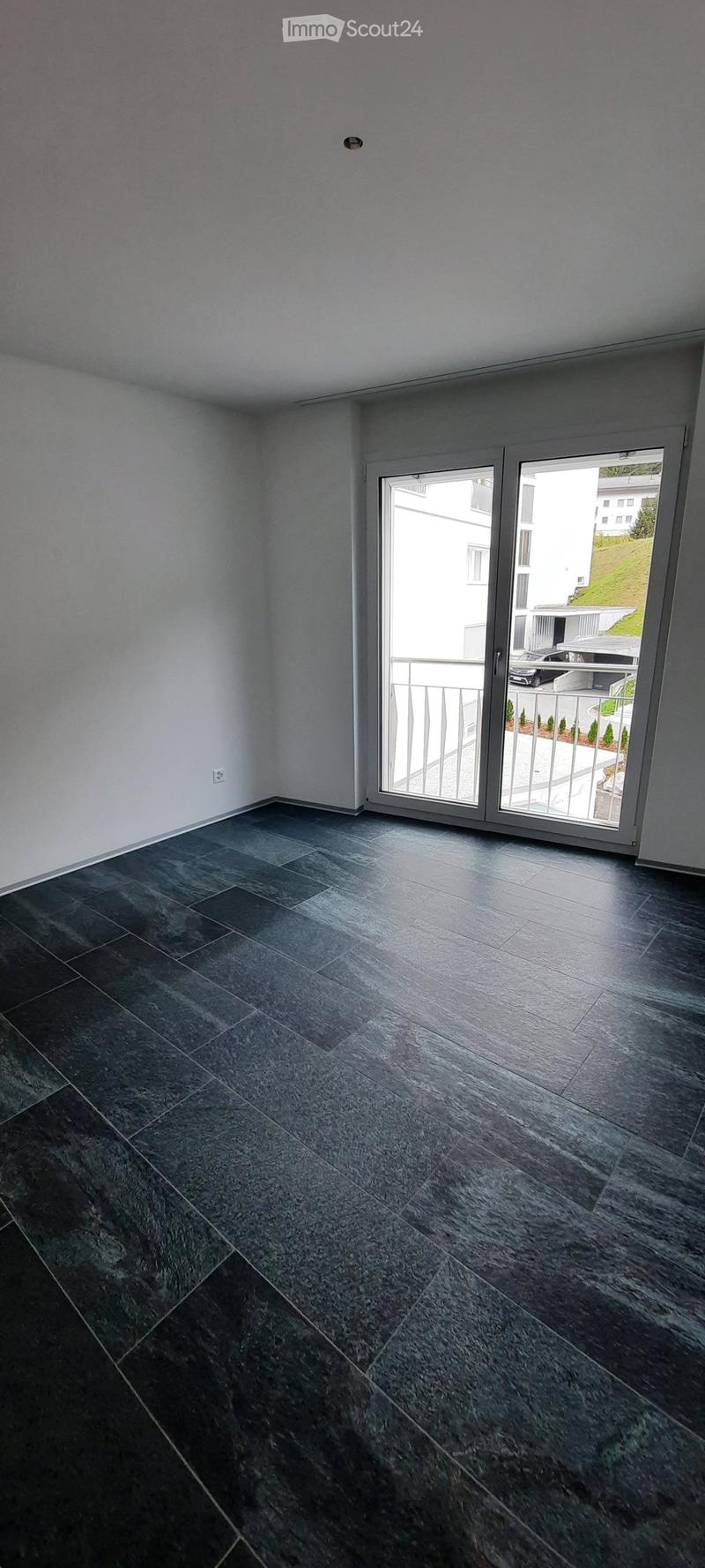 Empty room with dark grey tiled floor and white walls, featuring a large sliding glass door leading to a balcony.