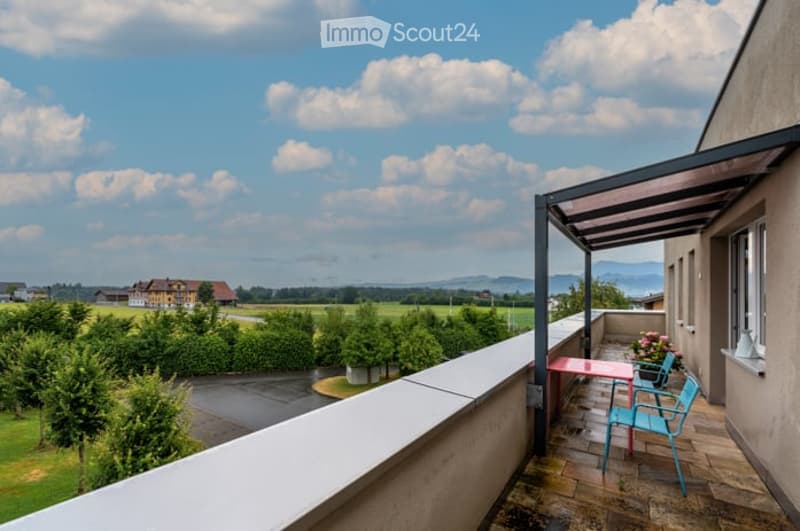 Balcony with a view of a green field and distant mountains, featuring a small seating area with a table and chairs under a canopy.