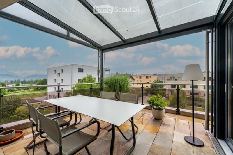 Modern balcony with a glass roof, white dining table, four chairs, potted plants, and a floor lamp, overlooking a scenic landscape with buildings and mountains.