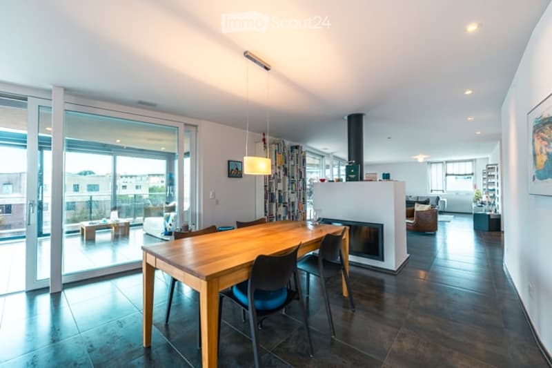 Modern open-plan living and dining area with large glass doors leading to a balcony, a central dining table with chairs, a fireplace, a sofa, and a bookshelf.