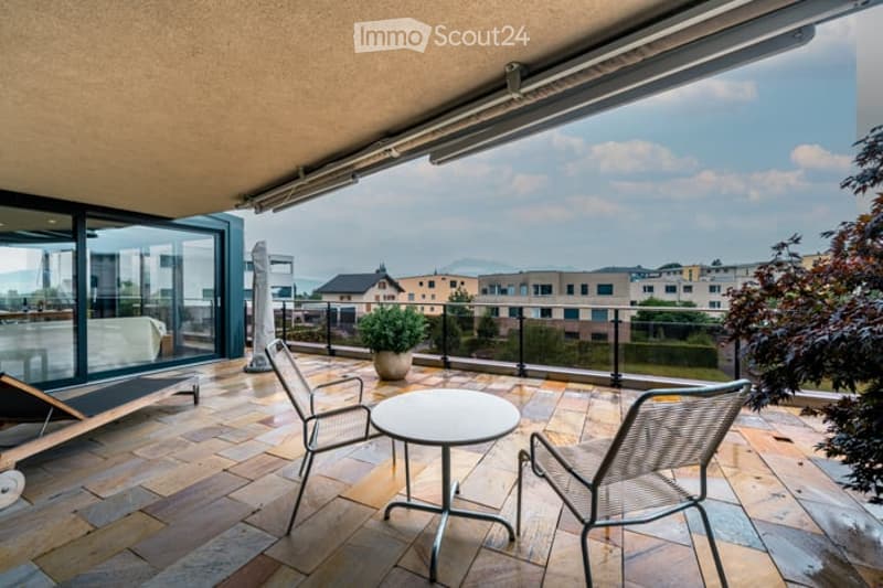 Balcony with tiled floor, glass railing, outdoor furniture including a table and chairs, potted plant, and a view of the city and mountains.