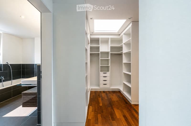 Modern bathroom with a bathtub, sink, and built-in wardrobe with shelves and drawers. The room features a wooden floor and a skylight above the wardrobe.