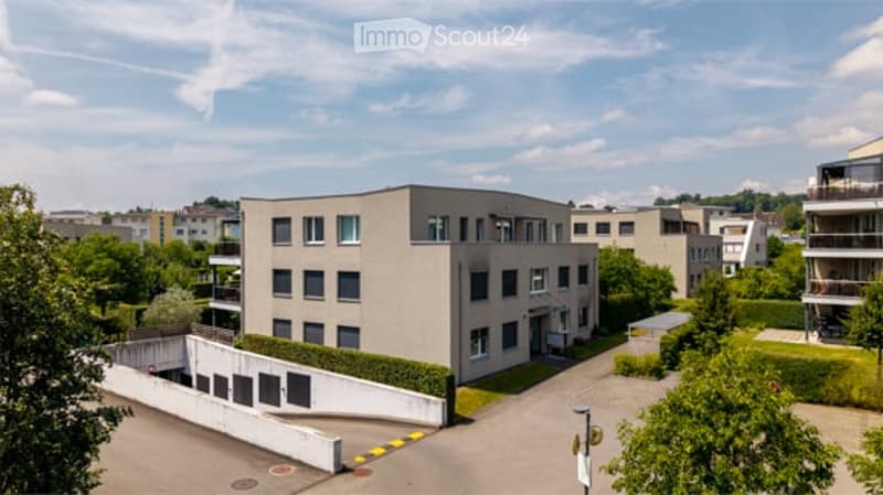 Modern multi-story building with a flat roof and rectangular design, surrounded by greenery and other residential structures.