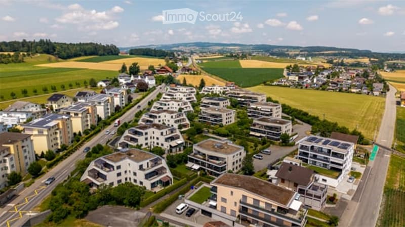 Aerial view of a residential area with multiple modern apartment buildings, surrounded by green fields and a road running through the neighborhood.