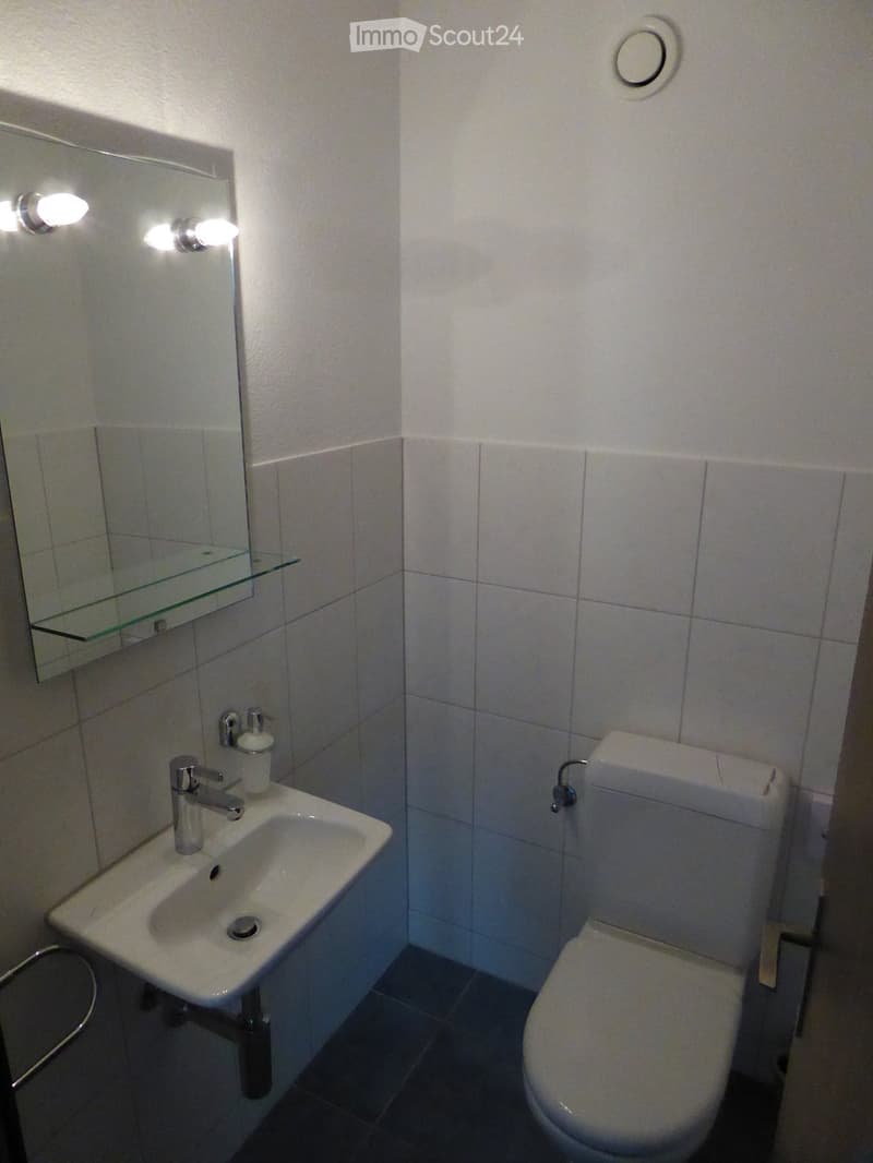 Modern bathroom with white tiled walls, a white sink with a chrome faucet, a toilet, a mirror with a glass shelf, and two wall-mounted light fixtures.