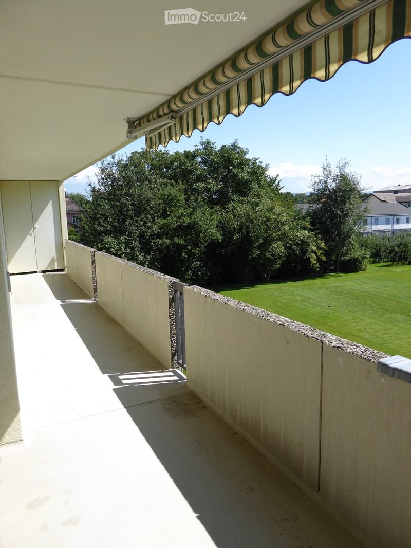 Balcony with a striped awning, low stone wall, and view of a green lawn and trees.