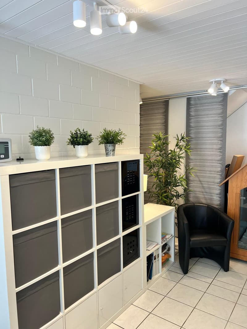 Modern interior with white brick wall, white ceiling lights, a black leather chair, a white shelf with books and potted plants, and a large screen mounted on the wall.