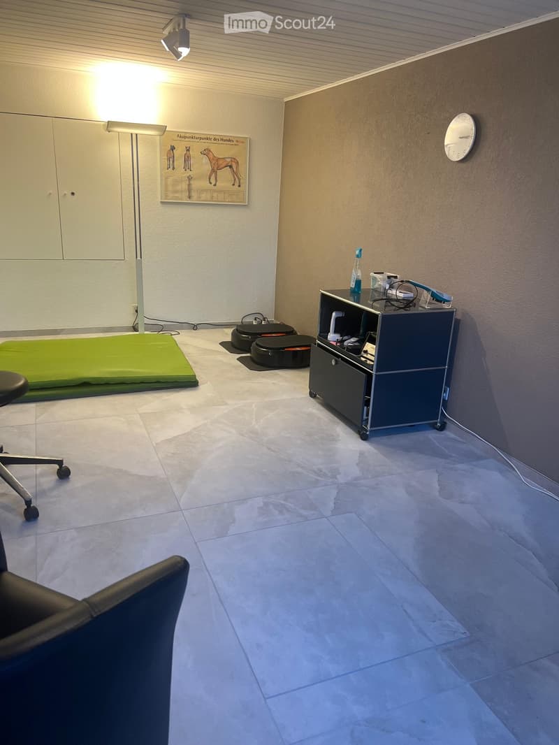 A clean, unfurnished room with tiled flooring, a green dog bed, a small cabinet with medical supplies, a poster on the wall, and a clock.