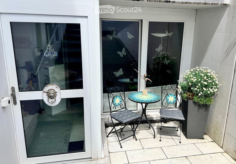Balcony with a small round mosaic table, two matching chairs, and a large flower pot with white flowers.