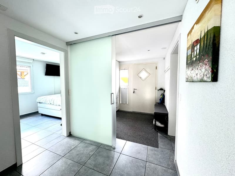 Hallway with tiled floor, glass sliding door, open doorway to bedroom with bed, TV, and window, and a small bench with shoes and umbrella.