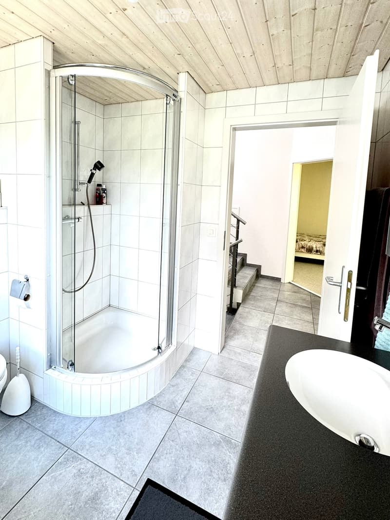Modern bathroom with a corner shower, bathtub, toilet, and a small sink area. The space features tiled walls and floors, a glass shower enclosure, and a small shelf with toiletries.