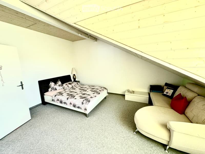Cozy attic bedroom with a slanted wooden ceiling, featuring a double bed with a black headboard and patterned bedding, a beige sectional sofa with red and black throw pillows, a small white side table, and a table lamp.