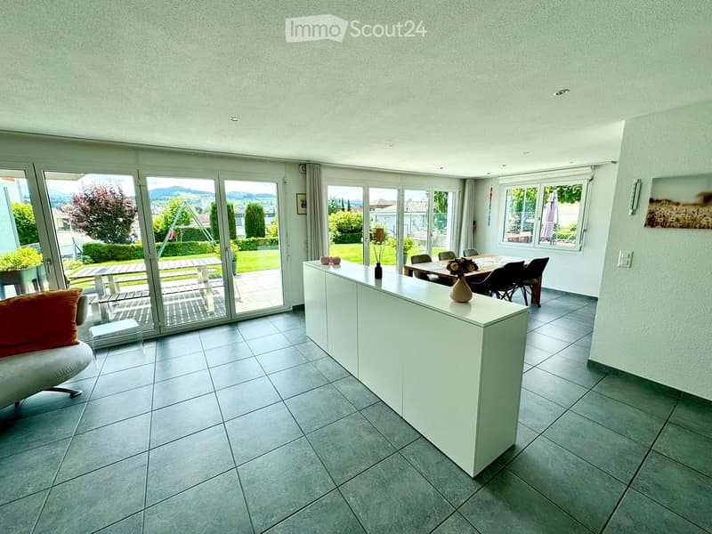 Modern living room with large windows and sliding glass doors leading to a patio, featuring a white sofa, dining table, and decorative vase.