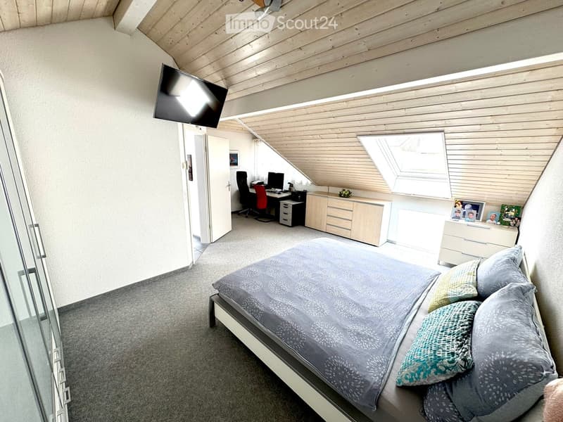 Bright bedroom with a large bed, desk area, and modern wooden ceiling. Features include a flat-screen TV mounted on the wall, a glass door, and a skylight window.